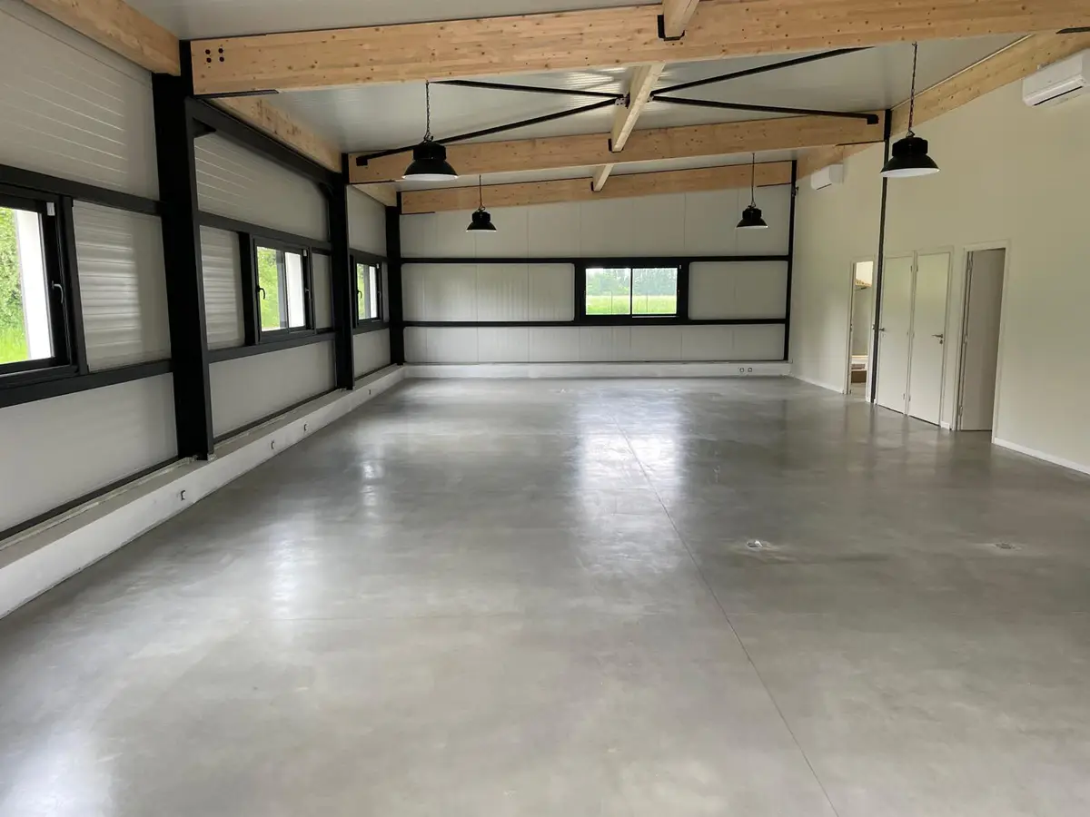 Grande salle vide au sol de béton lissé gris, poutres en bois clair et suspensions noires, propre après les travaux