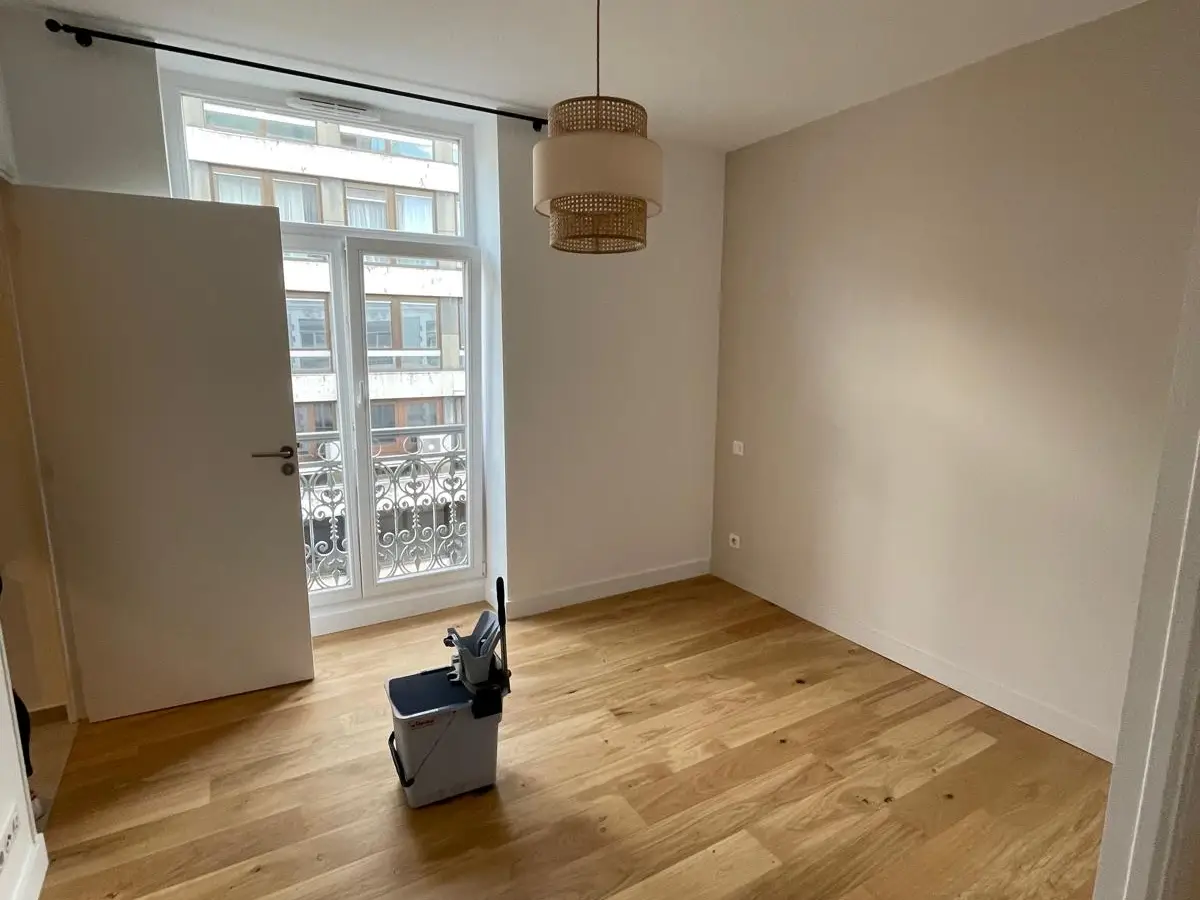 Pièce vide au parquet clair et à la porte-fenêtre sur balcon ouvragé, seau de lavage posé au milieu du sol
