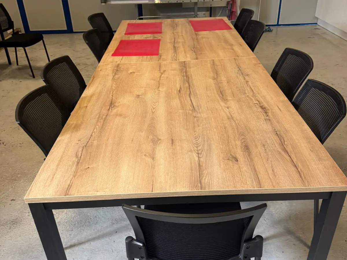 Grande table de réunion en bois clair entourée de chaises noires, dans une salle au sol de béton gris