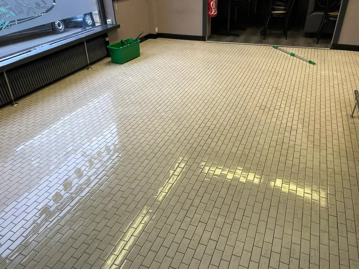Grande salle au sol carrelé beige encore humide après lavage, seau vert et manche télescopique posés près de la vitrine