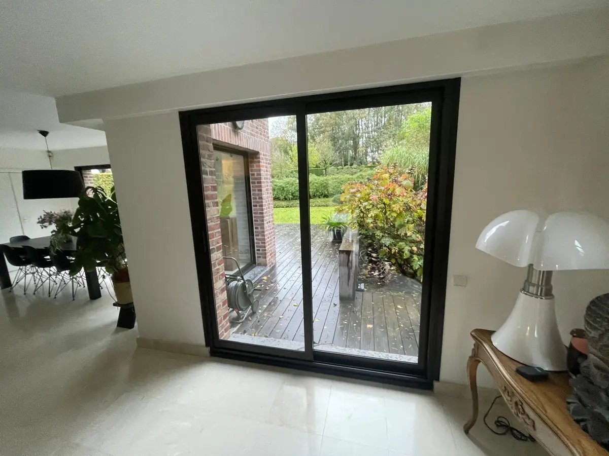 Baie vitrée coulissante à cadre noir vue de l’intérieur, avec une terrasse en bois et un jardin derrière