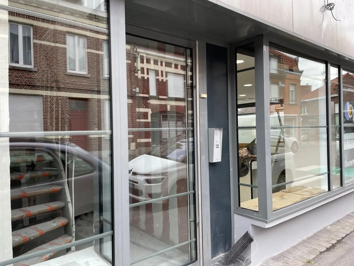 Vitrines et porte vitrée d’un local commercial en rue, cadres gris, façades en briques reflétées dans le verre